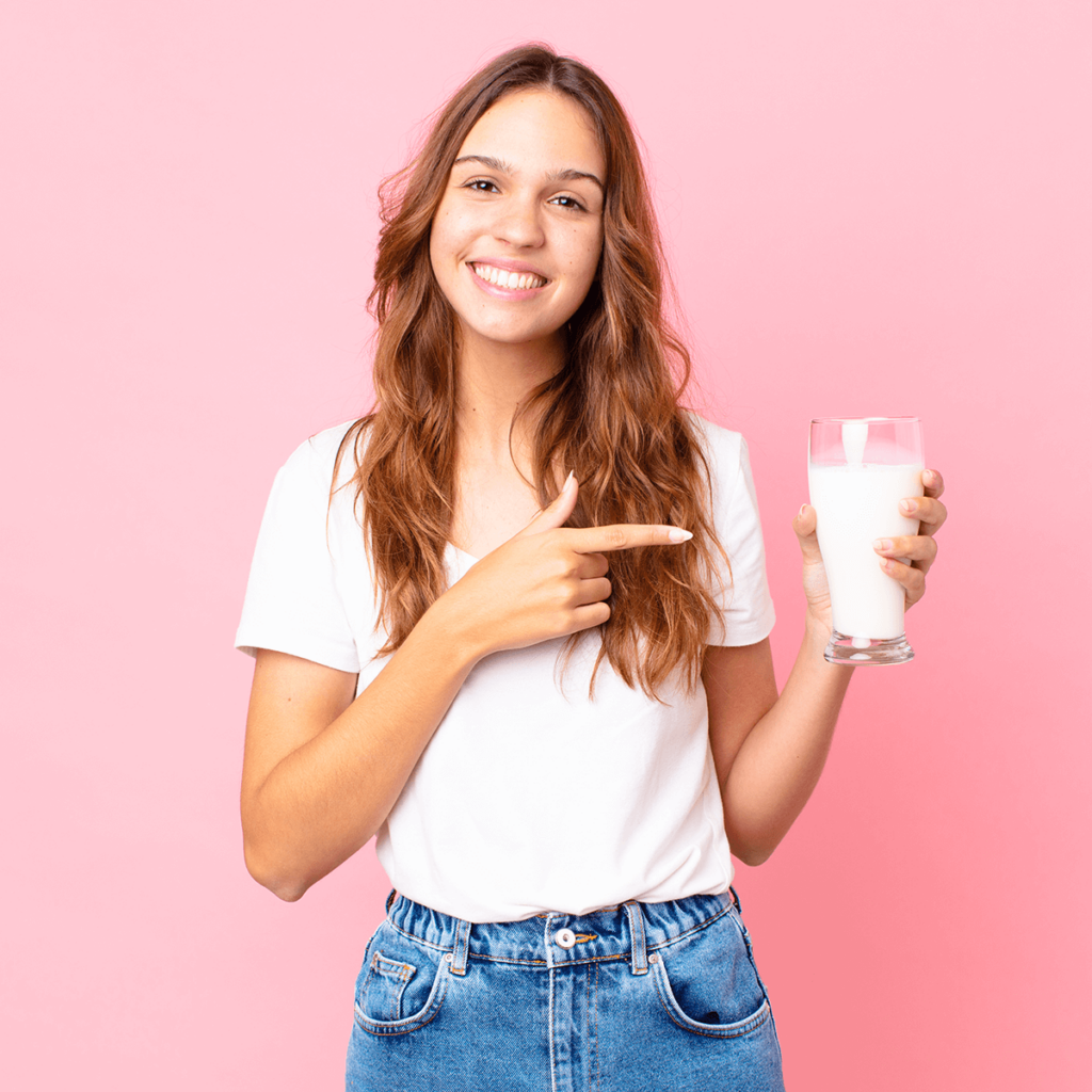 Eine junge Frau mit Laktoseintoleranz zeigt freudestrahlend auf ein Glas frischer Milch in ihrer Hand.